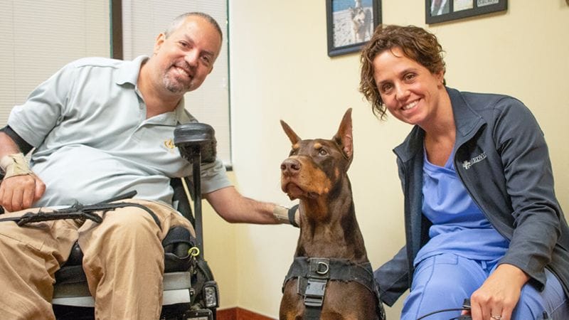 Brown Doberman service dog sits between the veterinary ophthalmologist and his owner’s wheelchair after his free eye examination